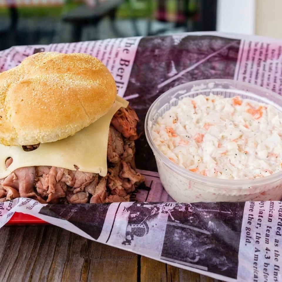 Cheesesteak sandwich with melted cheese and shredded beef beside a cup of creamy coleslaw on newspaper-lined tray.