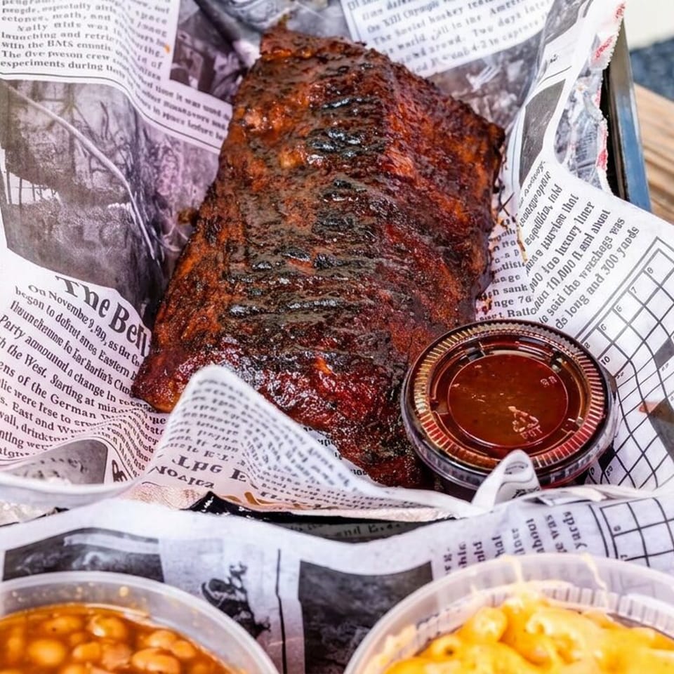A saucy grilled barbecue rib rack rests on newspaper with dipping sauce, baked beans, and macaroni and cheese on a wooden table.