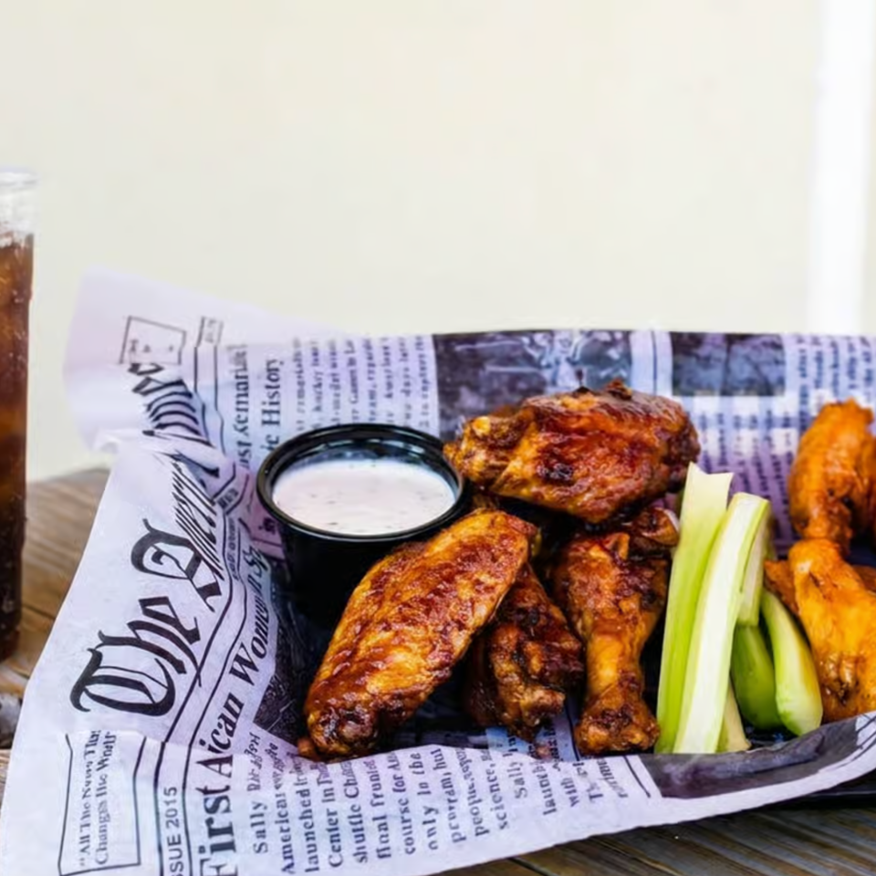 Basket of saucy chicken wings with celery, dipping sauce, and iced cola on a rustic wooden table.